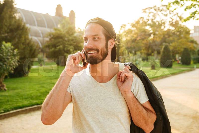 Portrait of muscular attractive guy wearing cap and white t-shirt holding leather jacket on shoulder and talking on cell phone ,during walk in green park, stock photo