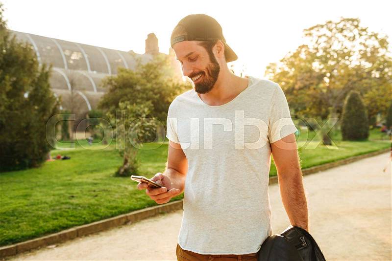 Image of muscular bearded guy in streetwear holding and chatting on mobile phone, while walking in green park, stock photo
