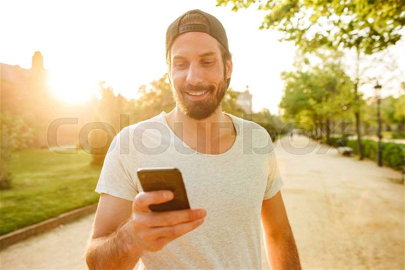Image of muscular macho man in casual wear using black smartphone for chatting, while walking in green park, stock photo