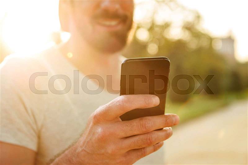 Cropped photo of cheerful caucasian man in casual wear smiling and using black smartphone for chatting, while walking in green park, stock photo