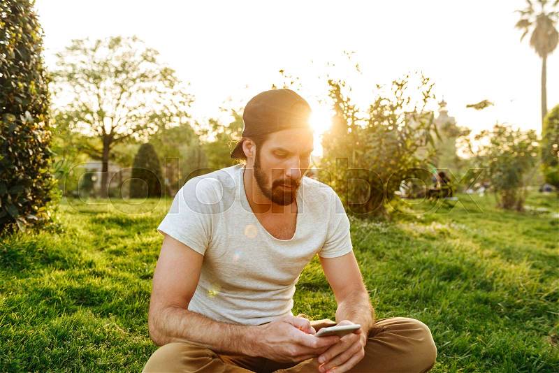 Image of caucasian handsome man in casual wear sitting on grass in green park, and using smartphone, stock photo