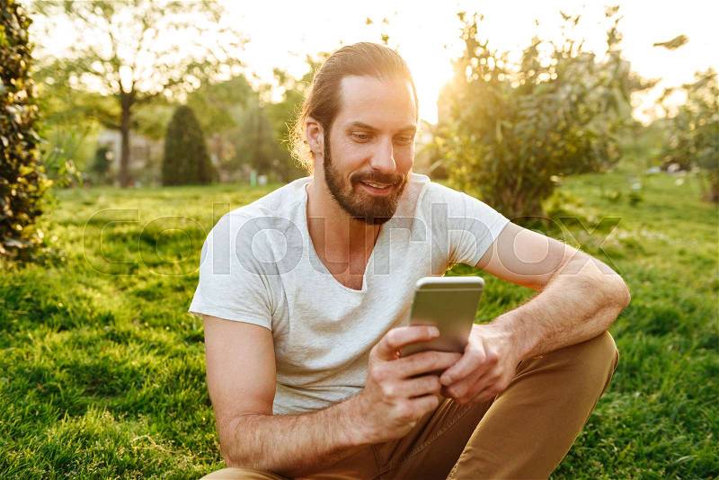 Image of caucasian handsome man in casual wear sitting on grass in green park, and using smartphone, stock photo