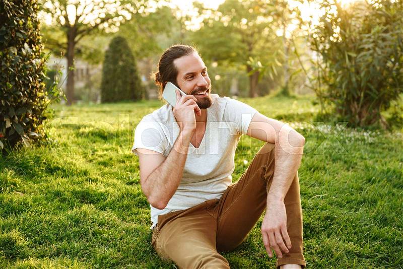 Picture of sociable young man in casual wear sitting on grass in green park, and speaking on smartphone with pleasure, stock photo