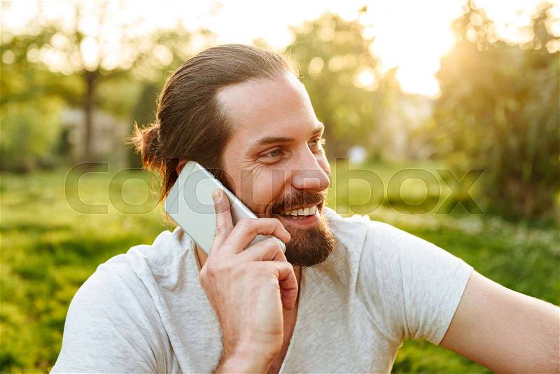 Picture closeup of positive young man in casual wear sitting on grass in green park, and chatting on mobile phone, stock photo