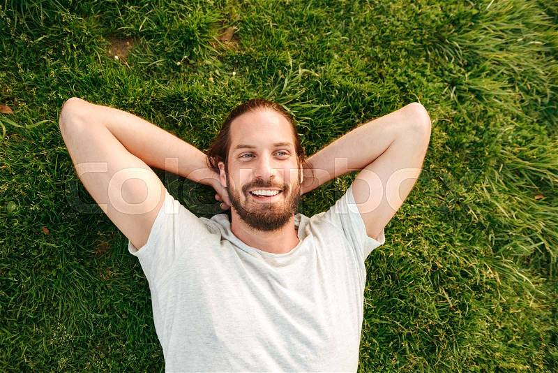 Photo from above of bearded handsome man 30s wearing white t-shirt lying on grass in green park, putting hands behind his back, stock photo