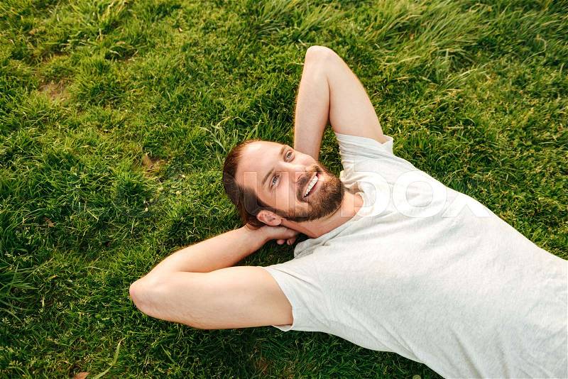 Photo from top of smiling handsome man wearing white t-shirt, spending leisure time in green park lying on grass and putting hands behind his back, stock photo