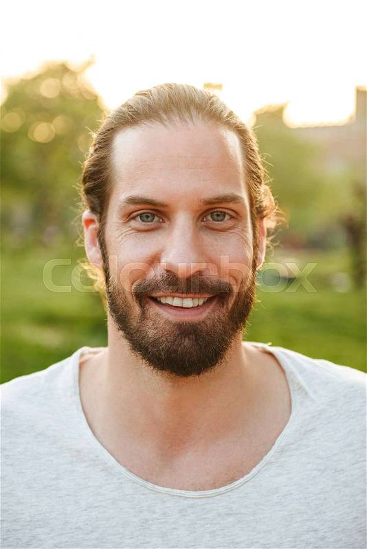 Profile closeup of handsome bearded man 30s with tied hair in white t-shirt smiling, while resting in green park, stock photo