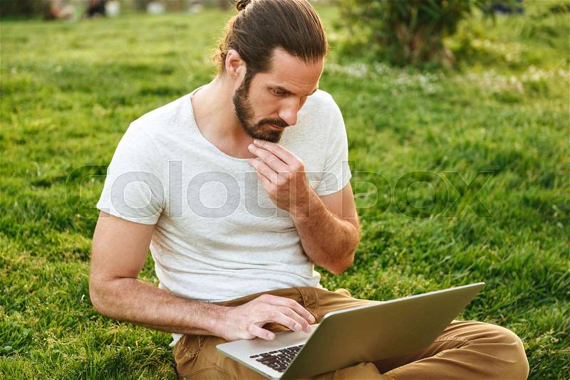Image of caucasian concentrated man in casual wear sitting on grass in green park, and working on silver laptop, stock photo