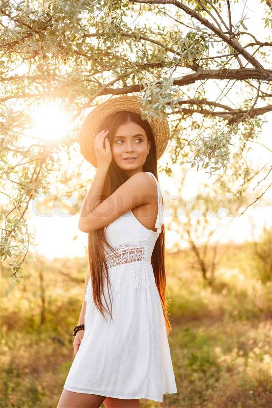 Photo of beautiful young woman with long dark hair wearing straw hat and white dress looking away, while standing under tree in nature during bright sunny day, stock photo