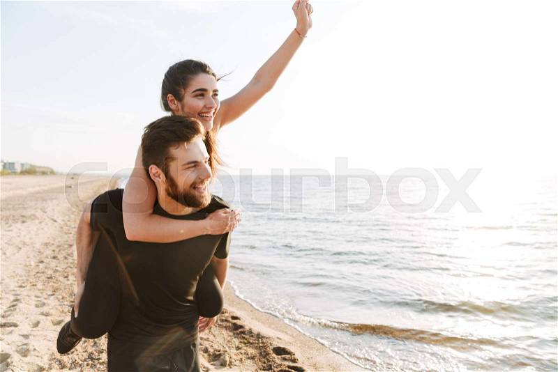 Happy young man carrying his girlfriend on his back while walking at the beach, stock photo