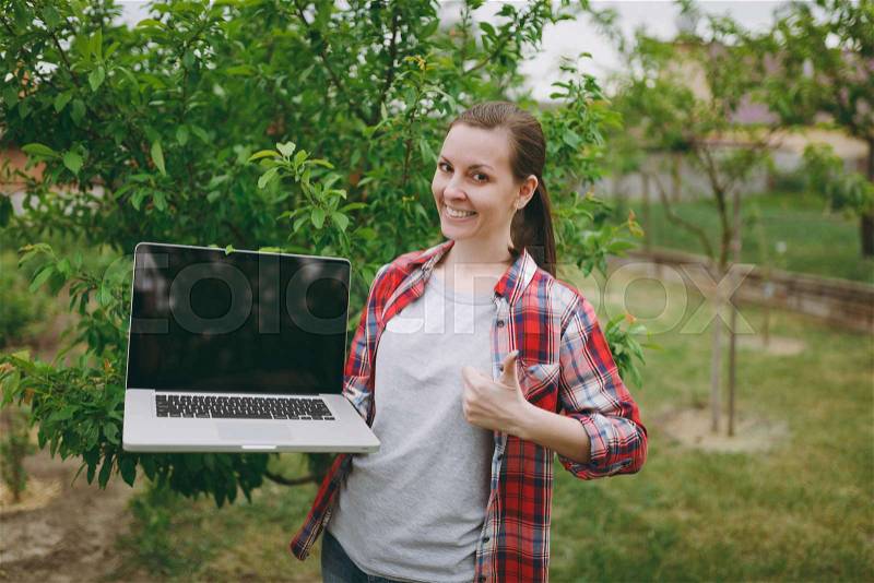 Young pretty woman in checkered shirt standing in garden near green tree, holding modern pc computer laptop with blank black empty screen. Copy space for advertisement. With place for text or photo, stock photo