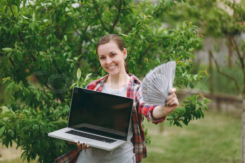 Woman standing in garden near green tree, holding bundle of dollars, cash money , modern pc computer laptop with blank black empty screen. Copy space for advertisement. With place for text or photo, stock photo