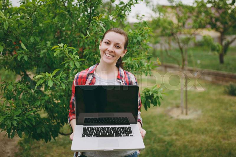 Young pretty woman in checkered shirt standing in garden near green tree, holding modern pc computer laptop with blank black empty screen. Copy space for advertisement. With place for text or photo, stock photo