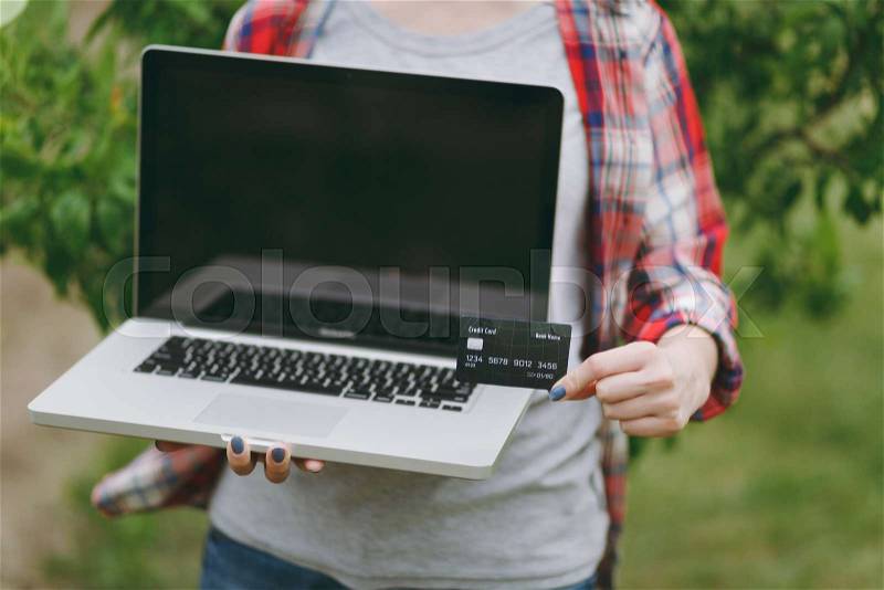 Close up cropped photo of woman standing in garden near green tree, holding credit card, modern pc computer laptop with blank black empty screen. Copy space for advertisement. With place for text, stock photo