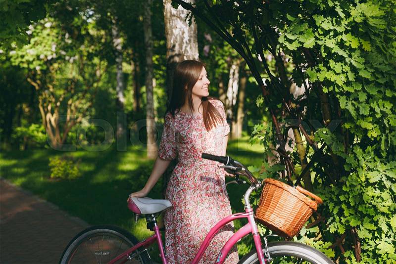 Portrait of trendy young woman in long pink floral dress stop to riding under oak arch on bike with basket for purchases, food or flowers outdoors, cute female recreation time in spring or summer park, stock photo