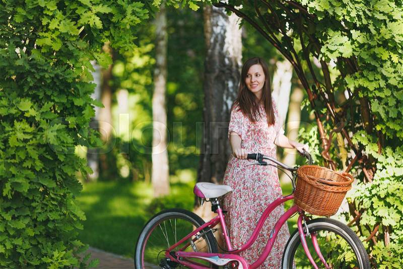 Portrait of trendy young woman in long pink floral dress stop to riding under oak arch on bike with basket for purchases, food or flowers outdoors, cute female recreation time in spring or summer park, stock photo