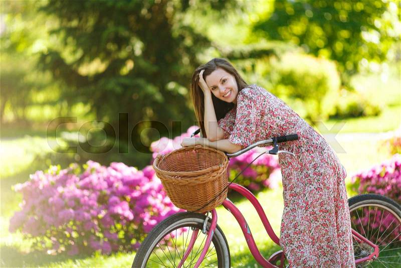 Portrait of trendy young woman in long pink floral dress stop to riding on vintage bike with basket for purchases on flowers background outdoors. Pretty female recreation time in spring or summer park, stock photo