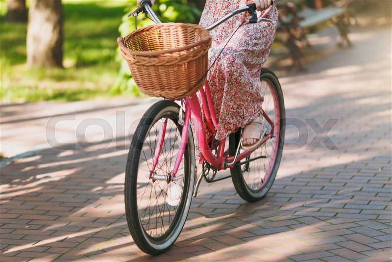 Close up cropped photo of young woman in long pink floral dress riding on vintage bike with basket for purchases, food or flowers outdoors, wheels, female recreation time in spring or summer park, stock photo