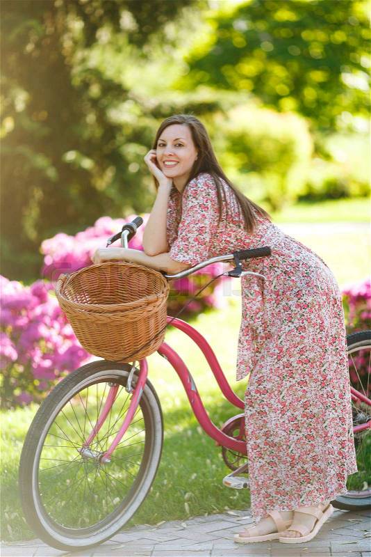 Portrait of smiling young woman in long pink floral dress stop to riding on vintage bike with basket for purchases on flowers background outdoors. Pretty female recreation time in spring, summer park, stock photo