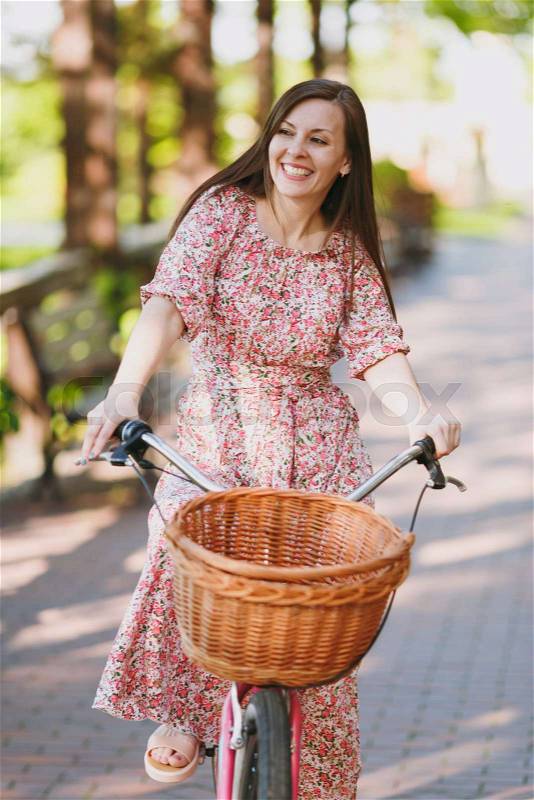 Portrait of trendy young woman in long pink floral dress riding on alley on vintage bike with basket for purchases, food or flowers outdoors, gorgeous female recreation time in spring or summer park, stock photo