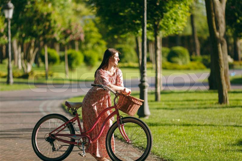 Portrait of trendy young woman in long pink floral dress stop to riding on vintage bike with basket for purchases, food or flowers outdoors, gorgeous female recreation time in spring or summer park, stock photo
