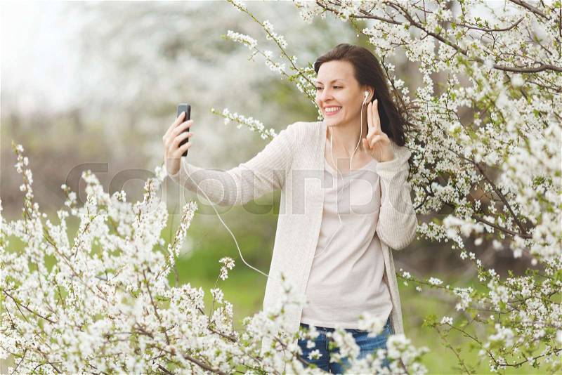 Young smiling woman in casual clothes with earphones listening music doing selfie on mobile phone showing victory sign in garden or park on blooming tree background. Spring flowers. Lifestyle concept, stock photo