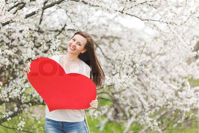 Young happy beautiful woman in light casual clothes holding big red heart standing in city garden or park on blooming tree background. Copy space for advertisement. Spring flowers. Lifestyle concept, stock photo