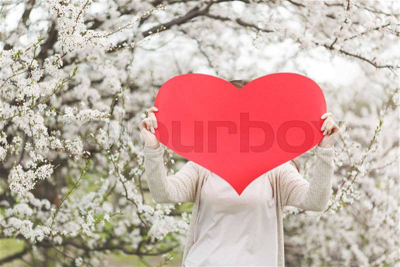 Young woman in casual clothes holding covering face with big red heart standing in city garden or park on blooming tree background. Copy space for advertisement. Spring flowers. Lifestyle concept, stock photo