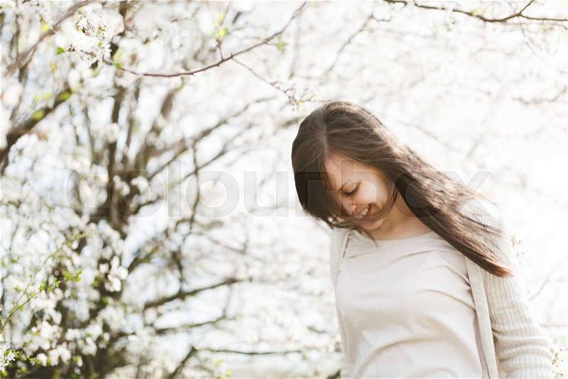 Young relaxed beautiful woman with lowered head and closed eyes in light casual clothes standing in city garden or park on blooming tree background. Spring nature, flowers. Lifestyle, leisure concept, stock photo