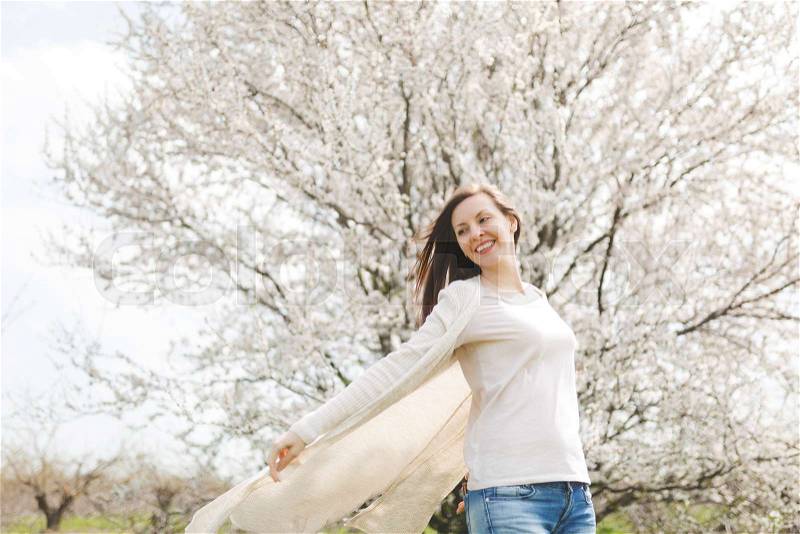 Young happy relaxed smiling beautiful woman in light casual clothes spreading hands standing in city garden or park on blooming tree background. Spring nature, flowers. Lifestyle, leisure concept, stock photo