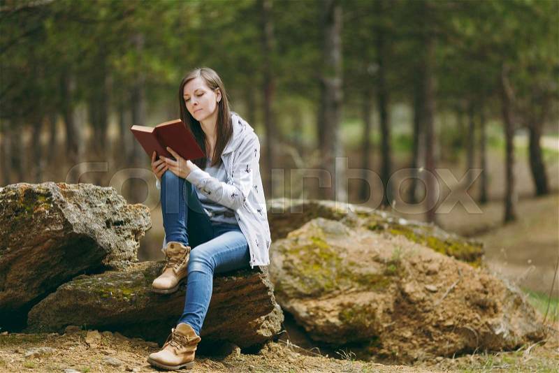 Young concentrated beautiful woman in casual clothes sitting on stone studying reading book in city park or forest on green blurred background. Student learning, education. Lifestyle, leisure concept, stock photo