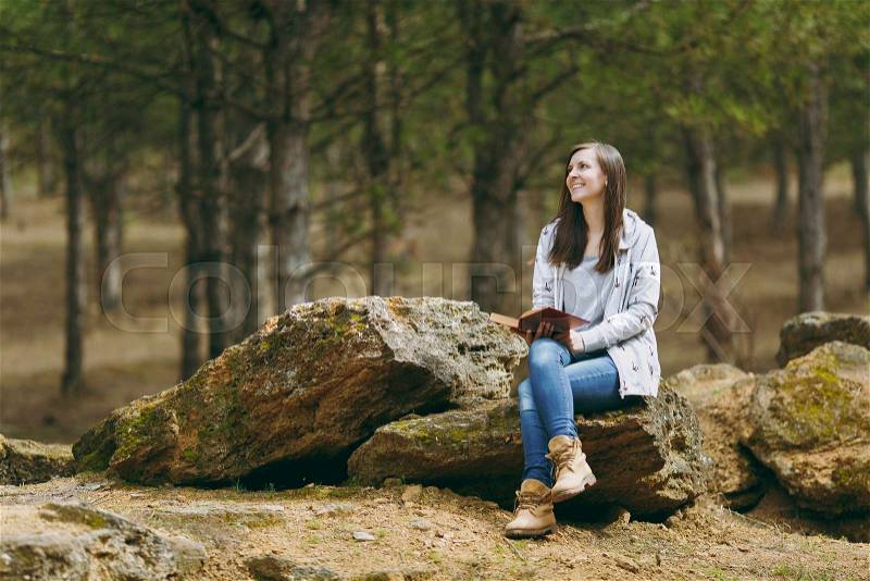 Young relaxing smiling beautiful woman in casual clothes sitting on stone studying reading book in big city park or forest on green background. Student learning, education. Lifestyle, leisure concept, stock photo