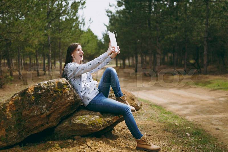 Young fun smiling woman in casual clothes with earphones sitting on stone using and doing selfie on tablet pc computer in city park or forest on green background. Student lifestyle, leisure concept, stock photo