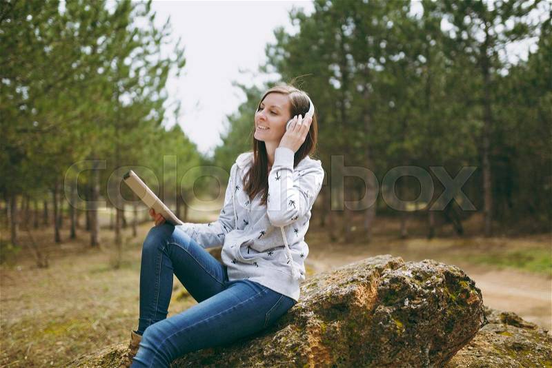 Young smiling woman in casual clothes sitting on stone listening music with headphones and tablet pc computer in city park or forest on green blurred background. Student lifestyle, leisure concept, stock photo