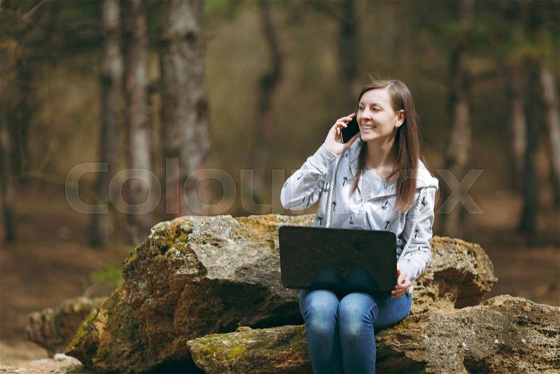 Young smiling business woman or student in casual clothes sitting on stone talking on mobile phone in city park or forest using laptop working outdoors on green background. Mobile Office concept, stock photo