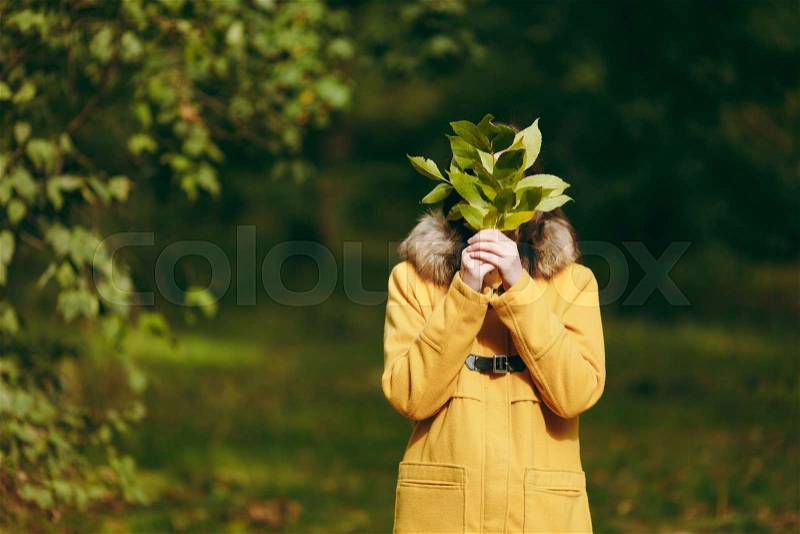 Beautiful happy caucasian young smiling brown-hair woman in yellow coat, jeans, boots in green forest. Fashion female model hiding behind fall leaves standing and walking in early autumn park outdoors, stock photo