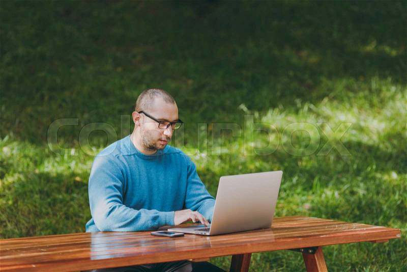 Young successful smart man businessman or student in casual blue shirt glasses sitting at table with mobile phone in city park using laptop working outdoors on green background. Mobile Office concept, stock photo