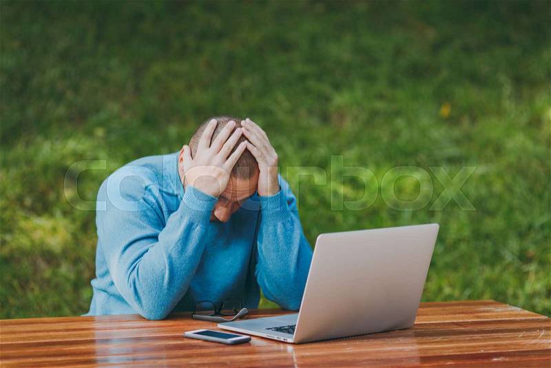 Young upset angry shocked man businessman or student in casual blue shirt, glasses sit at table in city park use laptop work outdoors put hands on head concerned about problems. Mobile Office concept, stock photo