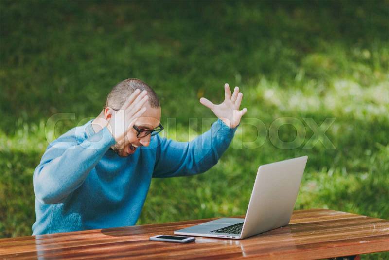 Young upset angry shocked man businessman or student in casual blue shirt glasses sitting at table in city park use laptop work outdoors throw up hands concerned about problems. Mobile Office concept, stock photo