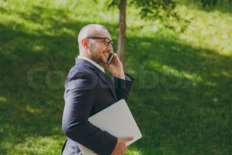 Successful smart businessman in white shirt, classic suit, glasses. Man stand with laptop pc computer, talk on mobile phone in city park outdoors on nature background. Side view, business concept, stock photo