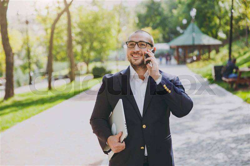 Successful smart businessman in white shirt, classic suit, glasses. Man stand with laptop pc computer, talk on mobile phone in city park outdoors on nature background. Mobile Office, business concept, stock photo
