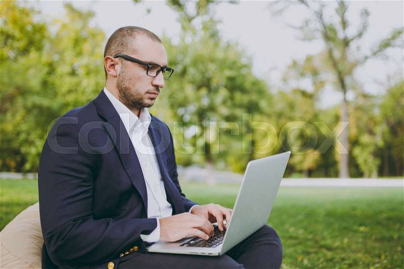 Young successful businessman in white shirt, classic suit, glasses. Man sit on soft pouf, working on laptop pc computer in city park on green lawn outdoors on nature. Mobile Office, business concept, stock photo