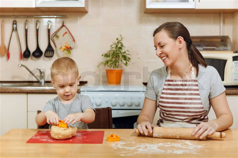 The little kid boy helps mother to cook Christmas ginger biscuit in light kitchen. Happy family mom 30-35 years and child 2-3 roll out dough and cut out cookies at home. Relationship and love concept, stock photo