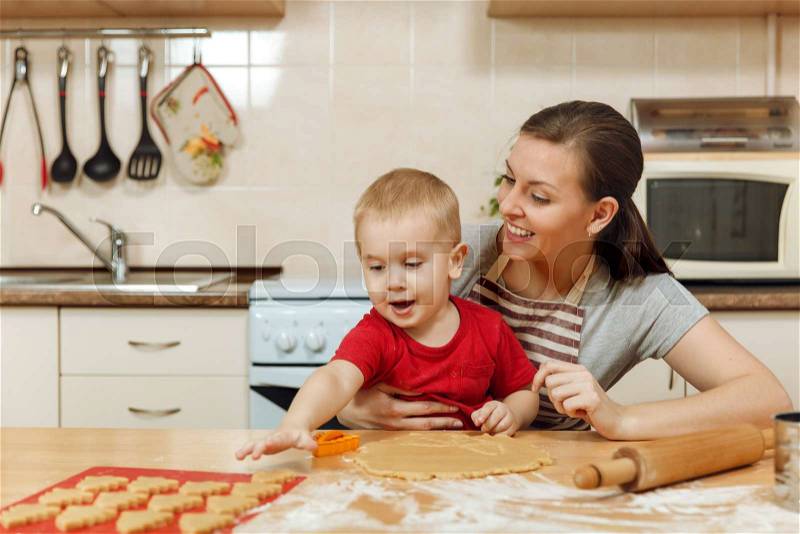 The little kid boy helps mother to cook Christmas ginger biscuit in light kitchen. Happy family mom 30-35 years and child 2-3 roll out dough and cut out cookies at home. Relationship and love concept, stock photo