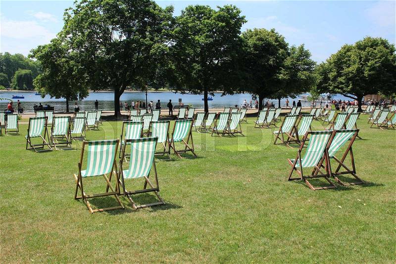 People and many deckchairs on the lawn in the sun along the Serpentine River in Hyde Park in the city London in England on a sunny day in spring, stock photo
