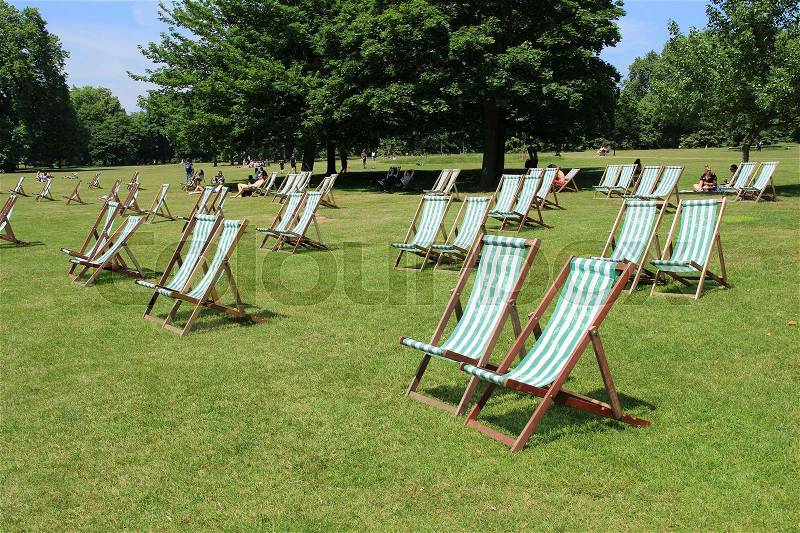 People and many deck chairs in the sun on the lawn in Hyde Park in the city London in England on a sunny day in spring, stock photo