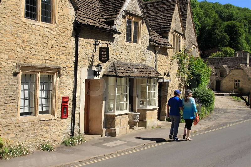 Couple, man and wife are walking along the old post office in one of the streets in the village Castle Combe in the Cotswolds in England on a sunny day in spring, stock photo