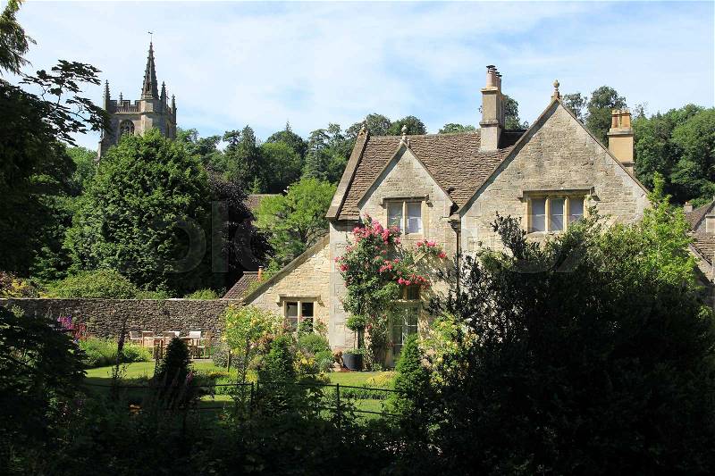The cottage with the beautiful garden and in the distance the church in the village Castle Combe in the Cotswolds in England on a sunny day in spring, stock photo