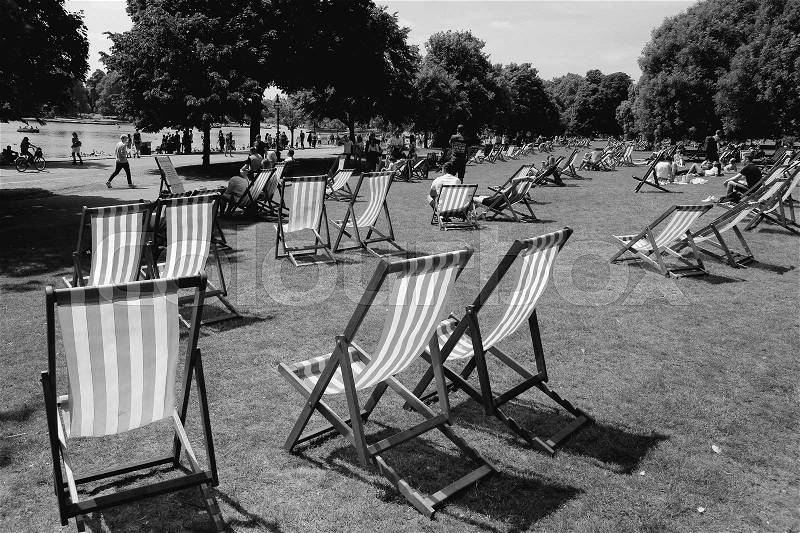 People and many deckchairs on the lawn in the sun along the Serpentine River in Hyde Park in the city London in England on a sunny day in spring in black and white, stock photo