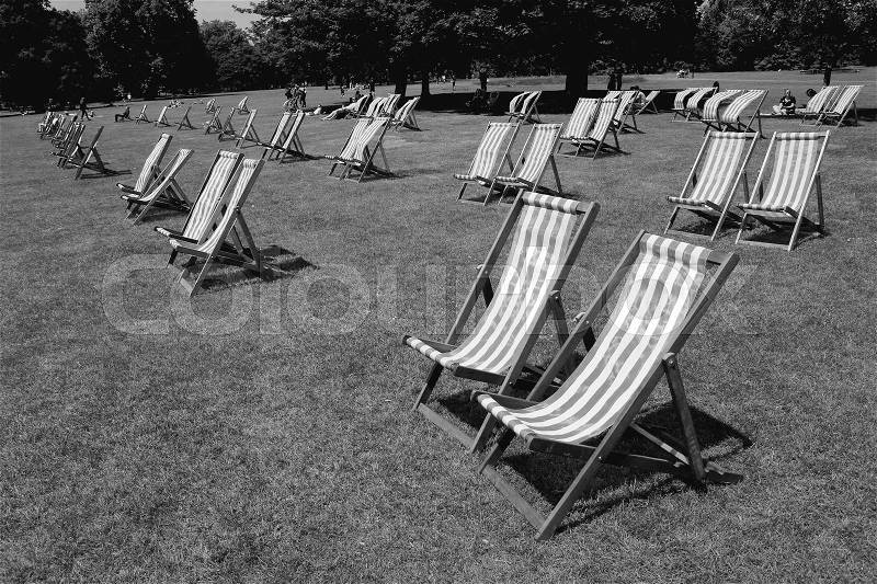 People and many deck chairs in the sun on the lawn in Hyde Park in the city London in England on a sunny day in spring in black and white, stock photo